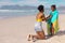 African american young mother wrapping happy daughter with towel while kneeling at beach against sky