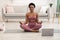 African Lady Meditating At Laptop Sitting In Lotus Position Indoors