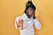 African man with dreadlocks wearing turtleneck sweater over yellow background covering eyes with hands and doing stop gesture with