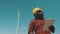 African man in hard hat standing with modern digital tablet against windmill power station making inspections of objects