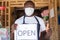 African small business owner wearing a face mask and holding an open sign in front of his shop does a thumbs up gesture