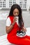 African stylish young woman in fashionable red suit drinks coffee on street. Cute black girl in youth trendy clothing with vintage