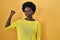 African young woman standing over yellow studio strong person showing arm muscle, confident and proud of power