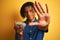 Afro american man with dreadlocks holding sunflowers seeds over isolated yellow background with open hand doing stop sign with