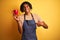 Afro barista man with dreadlocks drinking cup of coffee over  yellow background with surprise face pointing finger to