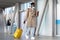 Afro millennial man walking with yellow suitcase in airport terminal, using mobile phone, wear face mask. Traveler african