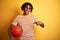 Afro sportsman with dreadlocks holding basketball ball over  yellow background with surprise face pointing finger to