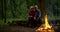 Aged loving pair is sitting on log in forest together and admiring flame of campfire