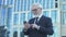 Aged male in formalwear scrolling phone standing near office center reading news