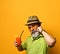 Aged man in hat, green shirt, sunglasses. Twisting mustache, holding glass of fresh squeezed juice, posing on orange background