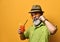 Aged man in hat, green shirt, sunglasses. Twisting mustache, holding glass of fresh squeezed juice, posing on orange background