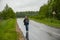 An aged woman walks along a wet highway that is in the woods