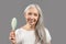 Aging gracefully. Charming senior lady with silky long hair holding brush and smiling on grey studio background