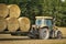 An agricultural tractor moves a bales of hay in a trailer on the field after harvesting grain crops.