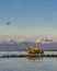 An airplane flies over a trebuchet fishing hut at sunset, against the snow-capped Alps, Marina di Pisa, Tuscany, Italy