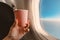 airplane passenger`s hand holds a paper Cup of tea against the blue sky in the window. Concept of the beginning of the journey