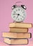 Alarm clock on top of piles of books on pink background.