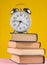 Alarm clock on top of piles of books on yellow background.