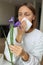 Allergic young woman holds iris flower, covers nose with paper tissue has runny nose, sneezes from flowers pollen at