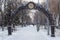 Alley with lanterns and benches under an iron arch with a clock. Beautiful urban side street with trees without foliage