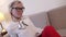 alluring blonde woman with glasses is flirting with camera, sitting on couch in apartment