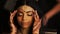 Alluring Indian bride holds her delicate fingers behind her temples while women get her hair ready