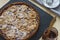 Almond cake with raspberry jam on a black graphite tray, a glass jar with a jam and a wooden spoon, a saucer with sugar powder.