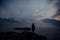 Alone human silhouette in white haze against the mountains and river. Thick fog in the evening twilight. Mysterious atmosphere.