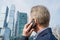 Alone middle-aged gray-haired businessman in a blue suit calling by phone standing opposite Moscow City skyscrapers.Low angle view