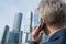 Alone middle-aged gray-haired businessman in a blue suit calling by phone standing opposite Moscow City skyscrapers.Low angle view