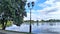 Along the shore of the lake is a tiled walkway. Nearby, there are trees and lamp posts with lights. On a sunny day, the water refl