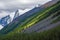 Alpine scenery with diagonal great snow mountain ridge coniferous forest and rocks under cloudy sky. Scenic sunny mountain