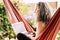 Alternative style caucasian people long grey white hair adult woman enjoy technology outdoor at home sit down on a red hammock