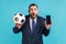 Amazed shocked man with beard in official style suit holding and showing black display smartphone and football ball, betting on