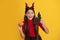 amazed teen girl wearing imp horns holding pumpkin and bat on yellow background, november