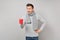 Amazed young man in gray sweater, scarf holding red cup of coffee or tea on grey background in studio. Healthy