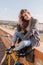 Amazing brown-haired girl in blue denim pants sitting on sky background in sunny morning. Glad white woman in gray coat