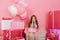 Amazing charming joyful little girl sitting with present suround big giftboxes, balloons on pink background. Happy