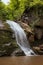 Amazing fresh summer waterfall in mountain with lush green forest, layered rocks overgrown moss and bright sunbeams on mountain.