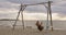 Amazing huge inflatable dinosaur is swinging on a swing on sea beach at sunset.