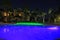 Amazing night landscape view. Blue pool water with green lights. Building with lighted windows and green plants on dark sky backgr