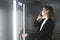 Ambitious office worker is pressing the button of the elevator and talking on her smartphone. Young employee is talking on her