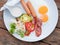 American breakfast with sunny side up fried eggs, bacon, sausage and vegetable salad on white dish on wooden table background