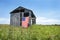 American flag hanging outdoors on side of old gray wooden barn on grass hill in countryside.