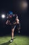 american football player holding ball and running on field under spotlights