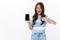 Amused cheerful young woman introduce smartphone app, hold mobile phone, pointing and looking display, smiling excited