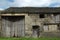 Ancient abandoned stone barn in a row of rural buildings with empty windows and wooden doors with the pavement overgrown with