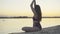 Angle view of relaxed slim woman sitting in lotus pose on sandy beach at sunset. Portrait of charming calm Caucasian