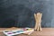 Angled view over a wooden desk with school supplies on it. Bucket with colored pencils, notebooks, paints. Back to school concept.