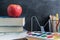 Angled view over a wooden desk with school supplies on it. Coloured pencils, books, calculator, tapes, paints and apple